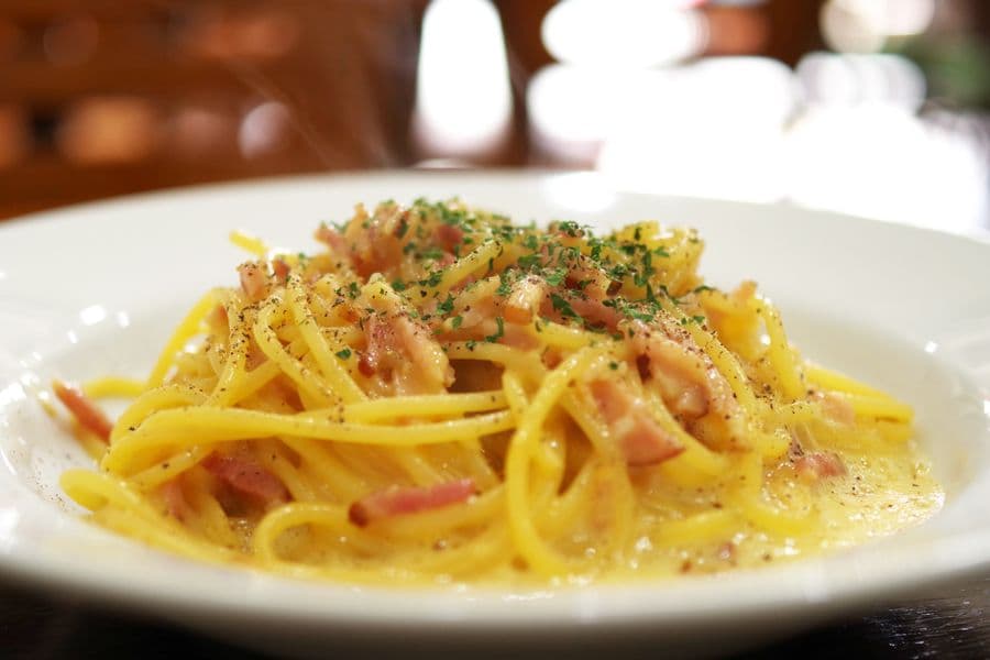 Karšti makaronai Carbonara su šonine, pabarstyti juodaisiais pipirais ir smulkintomis žolelėmis baltose lėkštėje.